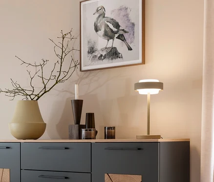 Ein dunkelgraues Sideboard mit heller Holzplatte trägt eine Vase mit Ästen, Kerzenhalter und eine leuchtende Tischlampe. Darüber hängt ein gerahmtes Vogelbild an einer beigen Wand.
