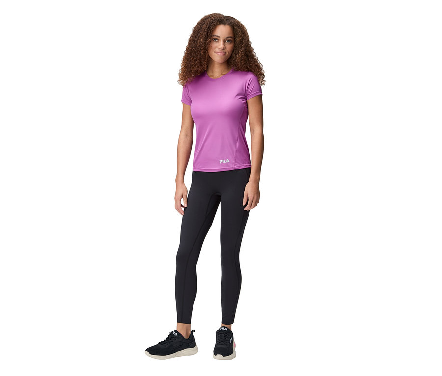 Eine Frau mit lockigem Haar trägt ein lilafarbenes FILA Damen-Funktionsshirt, schwarze Leggings und schwarze Turnschuhe.