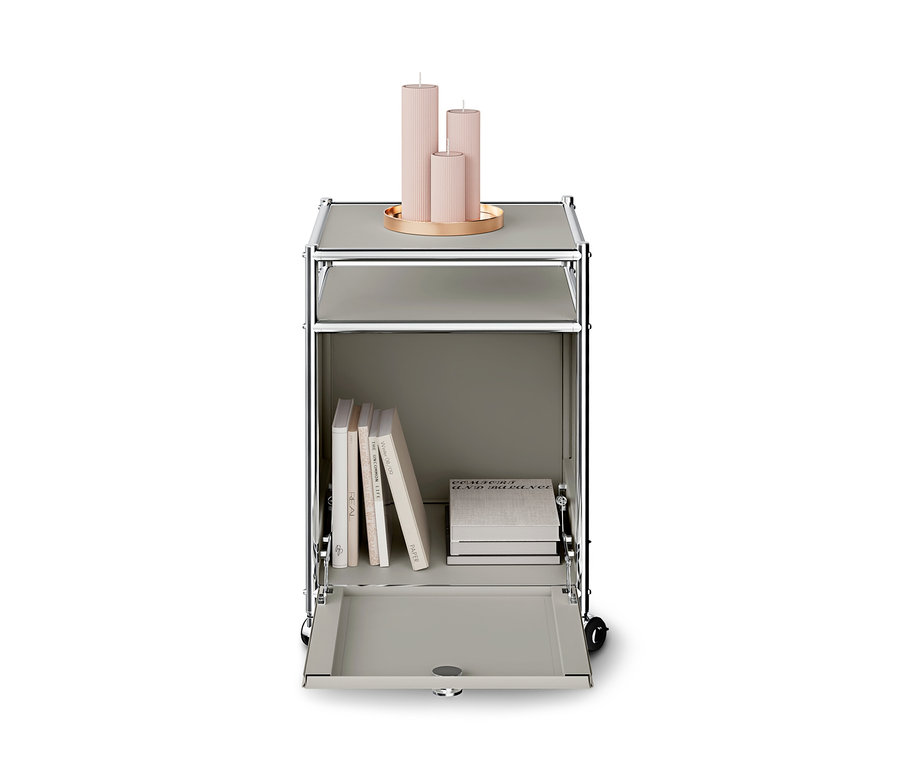 Grauer modularer Rollcontainer mit Chromrahmen. Obenauf steht ein goldenes Tablett mit drei gerippten, rosafarbenen Kerzen. Das untere Fach ist offen und zeigt mehrere Bücher, teils stehend, teils gestapelt.