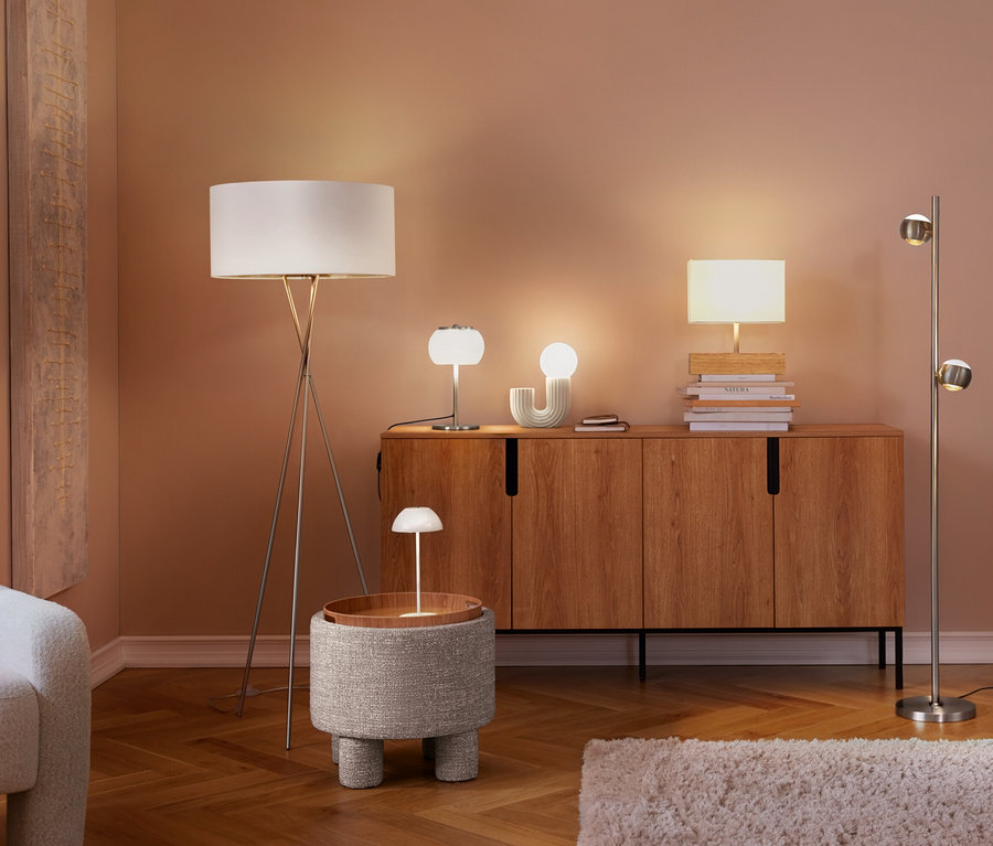 Ein Wohnzimmer mit mehreren eingeschalteten Lampen. Eine weiße LED-Akku-Tischleuchte steht auf einem runden Beistelltisch, daneben ein Sessel und ein Holzschrank mit weiteren Lampen.
