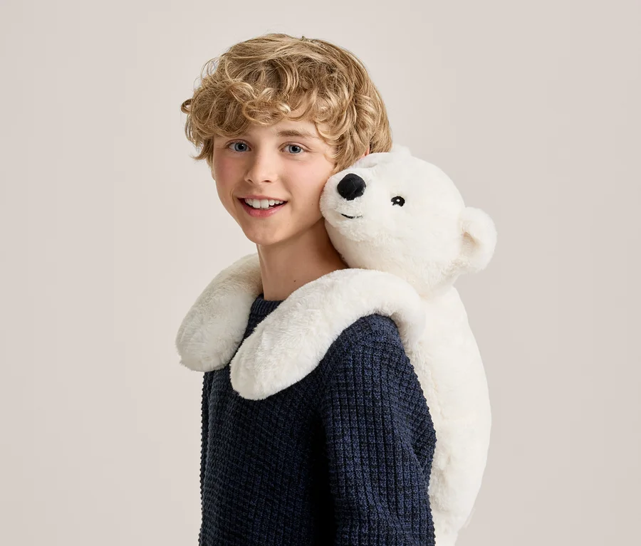 Lächelnder Junge mit lockigem blondem Haar in einem dunkelblauen Pullover, der von einem weißen Eisbären-Plüschtier von hinten umarmt wird, das über seinen Schultern liegt.