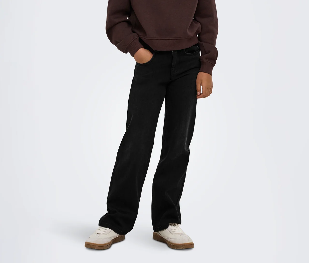 Person trägt ein braunes Sweatshirt, schwarze weite Jeans mit einer Hand in der Tasche und hellbeige Sneaker.