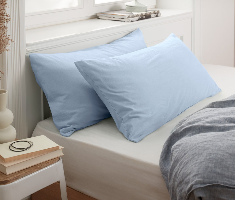 Zwei blaue Jersey-Kopfkissenbezüge (je ca. 80 x 40 cm) liegen auf einem Bett neben einem Nachttisch mit Vase und Büchern.