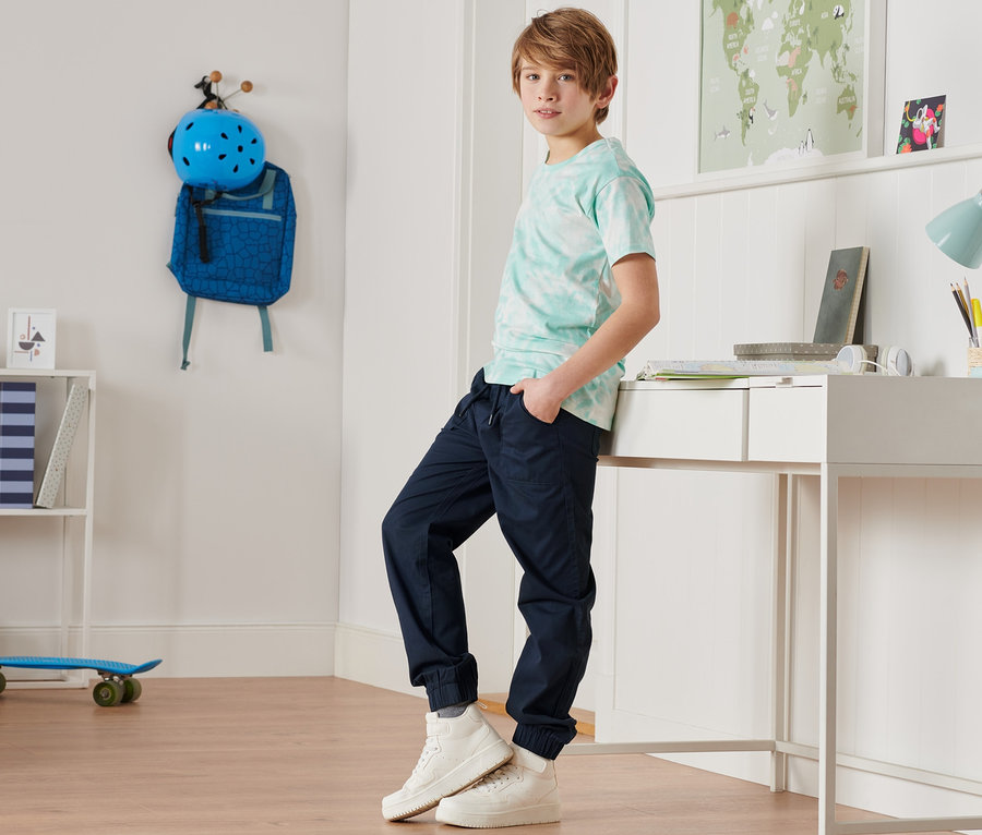 Ein Junge posiert in blauen Joggpants aus Bio-Baumwolle und einem türkisfarbenen T-Shirt in einem Zimmer.