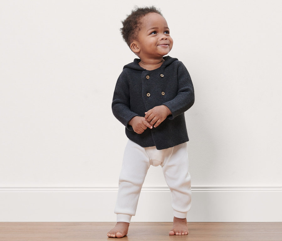 Ein lächelndes Baby steht barfuß auf dem Boden und trägt eine dunkelgrau melierte Baby-Strickjacke.