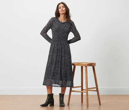 Frau posiert in einem gepunkteten Mesh-Kleid neben einem Hocker.