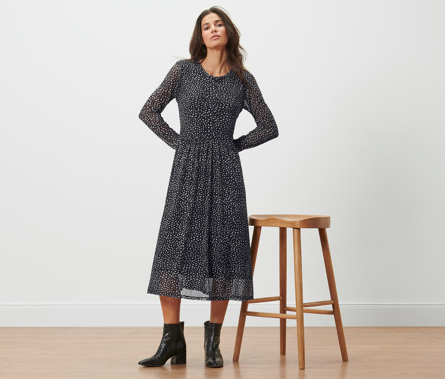 Frau posiert in einem gepunkteten Mesh-Kleid neben einem Hocker.