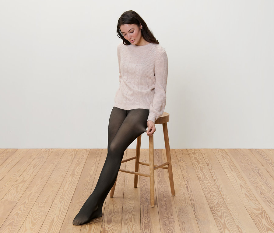 Eine Frau sitzt auf einem Hocker und zieht eine gefütterte Feinstrumpfhose in transparenter Optik an.