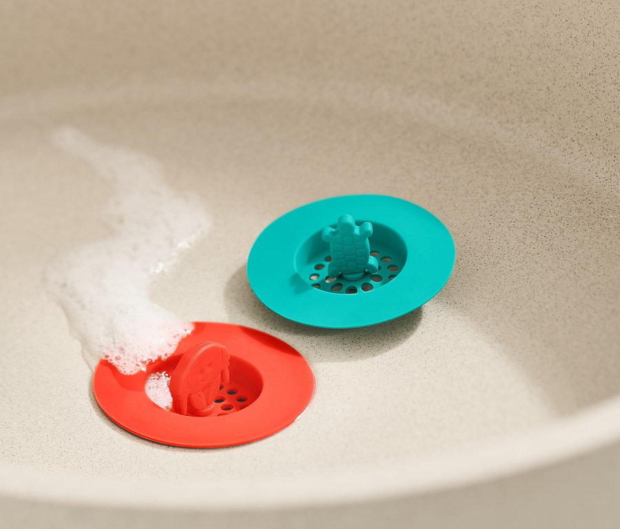 Im Waschbecken liegen zwei Abflusssiebe aus Silikon in Rot und Türkis. Unter dem roten Sieb fließt Wasser.