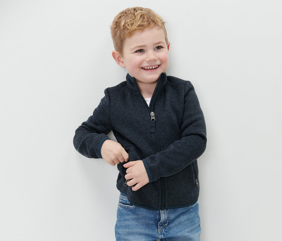 Lächelnder Junge steht an einer Wand und trägt eine Kinder-Strickfleecejacke und blaue Jeans.