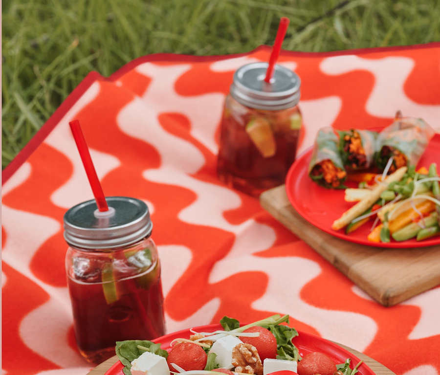 Auf einer XL-Picknickdecke liegen ein Teller mit Salat, Picknick-Besteck und Gläser mit Getränken und Kunststoff-Trinkhalmen.