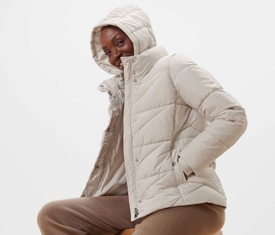 Frau sitzt auf einem Hocker und trägt eine Steppjacke mit Kapuze.