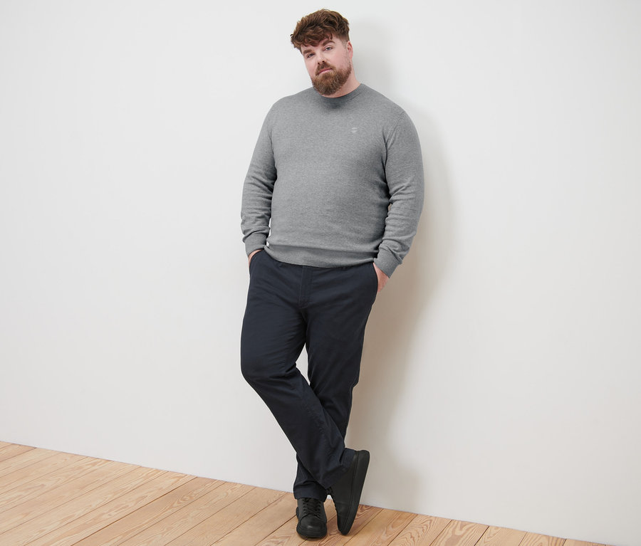 Mann lehnt an Wand, trägt grauen Pullover mit Rundhalsausschnitt und marineblaue Chino.