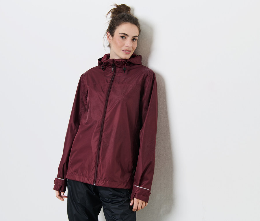 Eine Frau lehnt an einer Wand und trägt eine weinrote Regenjacke.