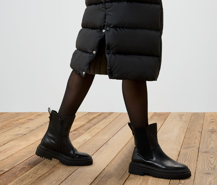 Teilansicht einer Person mit einem schwarzen Daunenrock, einer schwarzen Strumpfhose und schwarzen Lederstiefeln auf einem Holzboden.