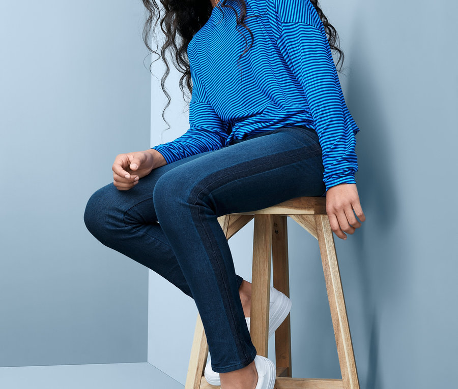 Junge Frau sitzt auf einem Holzhocker und trägt blaue Stretch-Jeans und ein gestreiftes Oberteil.