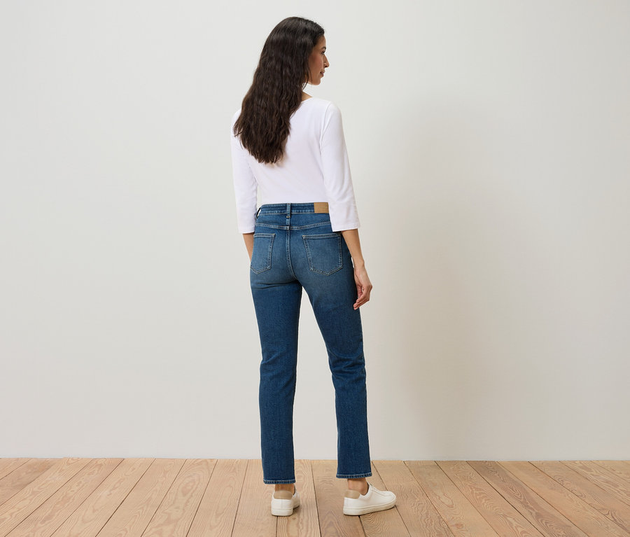 Frau steht mit dem Rücken zur Kamera und trägt blaue Jeans, ein weißes Hemd und weiße Turnschuhe.
