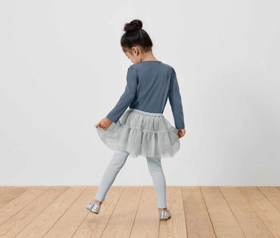 Mädchen in grauem Langarmshirt, silbernem Tutu-Rock und grauen Leggings. Hält den Rock.