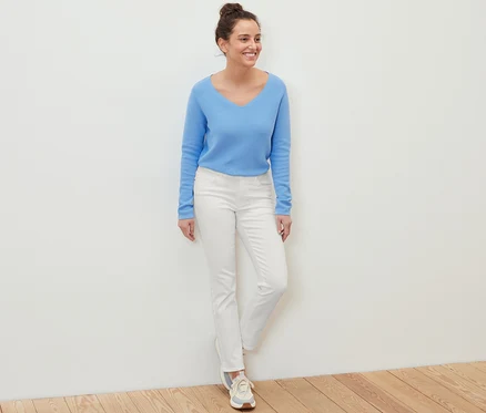 Eine Frau steht und lächelt, sie trägt einen blauen Pullover und weiße Jeans ‒ Fit »Kira«.