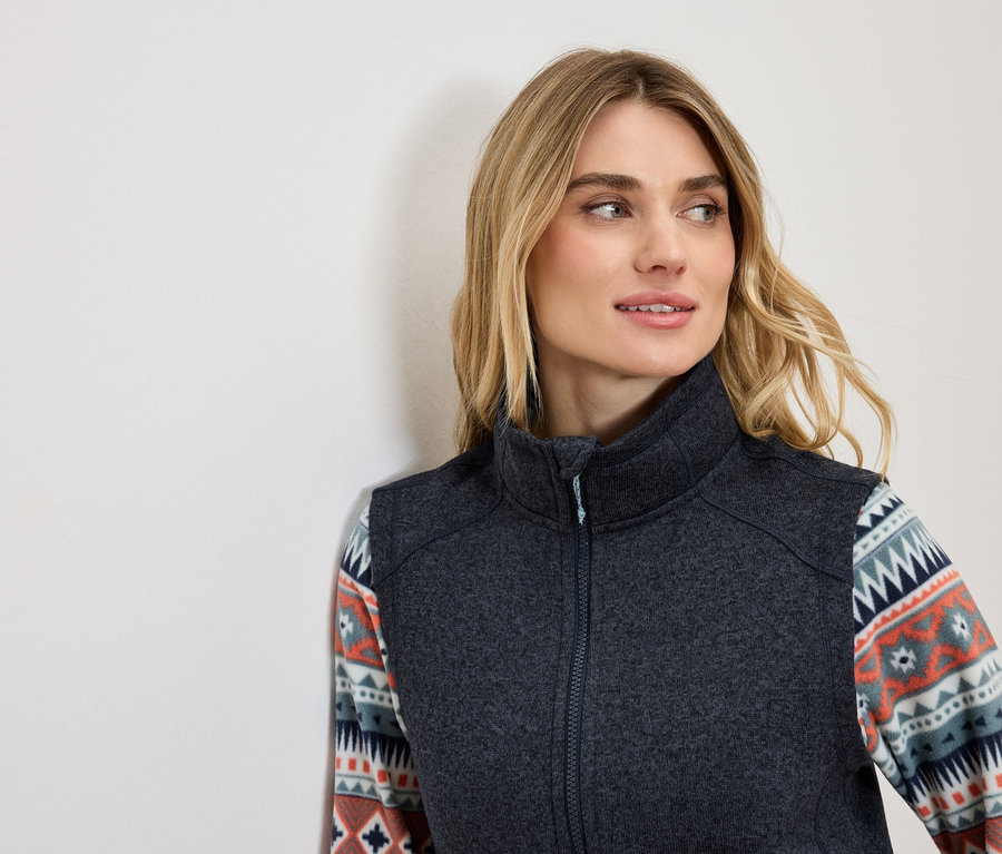 Blonde Frau in dunkelgrauer Weste mit dekorativem Muster auf den Ärmeln, die nach links schaut.