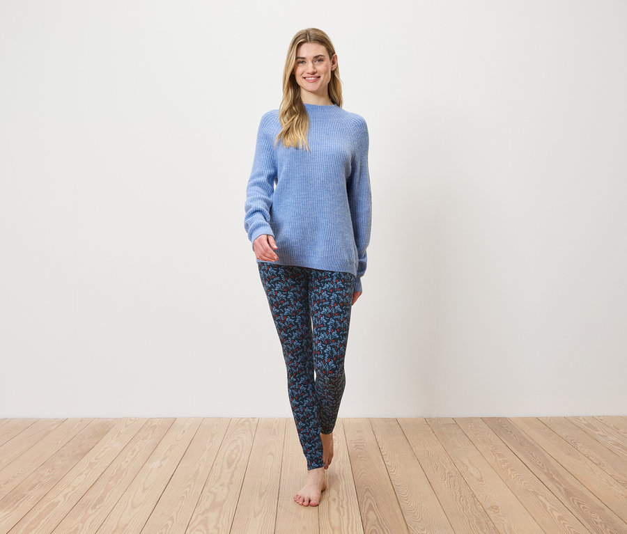 Eine Frau geht barfuß auf einem Holzboden und trägt einen blauen Pullover und gemusterte Leggings.