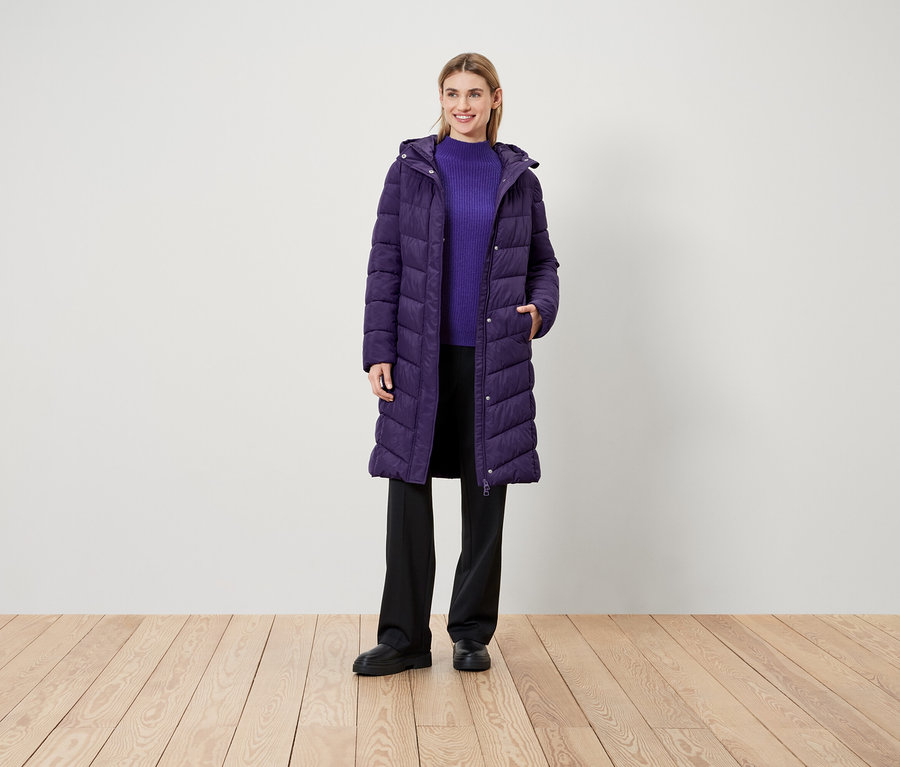 Eine Frau steht auf einem Holzboden und trägt einen lila Mantel, einen lila Pullover und eine schwarze Hose.