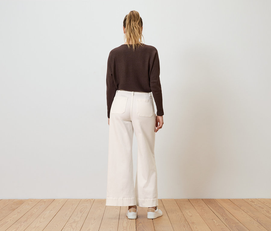 Eine Frau mit Pferdeschwanz steht mit dem Rücken zur Kamera und trägt einen braunen Pullover, weite, cremefarbene Hosen und weiße Turnschuhe.