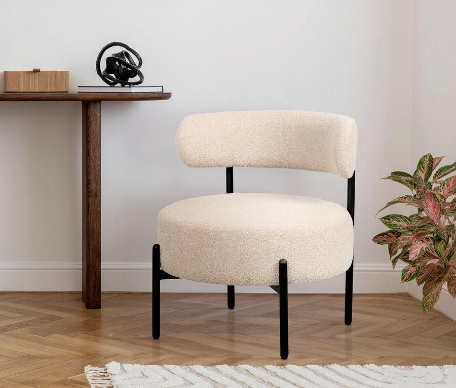 Ein cremefarbener Stuhl mit schwarzem Gestell steht auf einem gemusterten Teppich. Ein Holztisch mit Deko steht daneben.