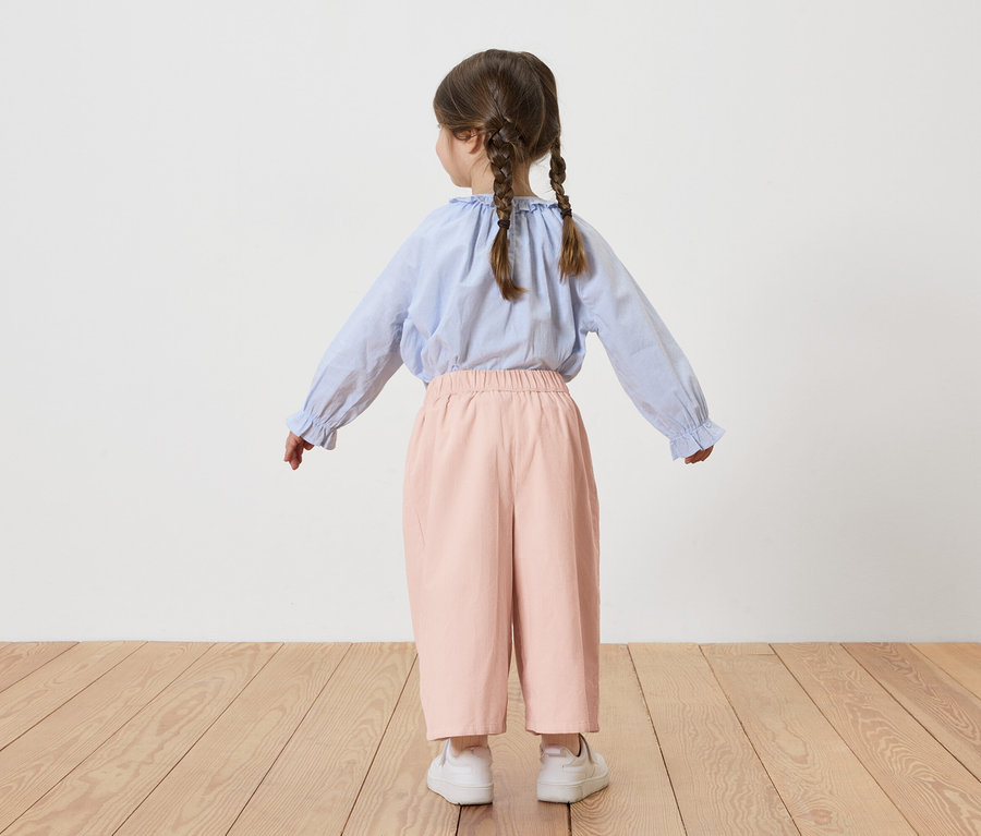 Ein kleines Mädchen mit Zöpfen, von hinten gesehen, trägt eine hellblaue Bluse und eine rosafarbene Hose, während es auf einem Holzboden steht.