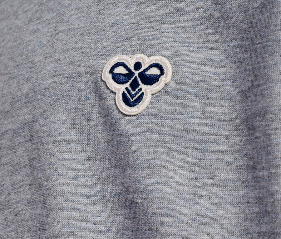 Detailansicht eines hellgrauen Stoffes mit einem kleinen, aufgenähten, weißen und blauen Hummel-Logo.