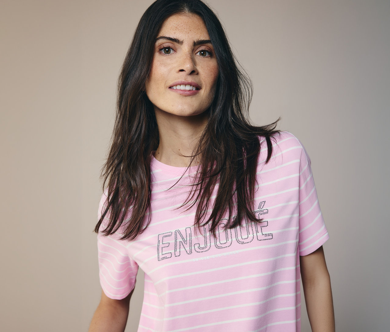 Frau mit langen, dunklen Haaren trägt ein CECIL Shirt mit rosa-weißen Streifen und dem Wort 'ENJOUE'.