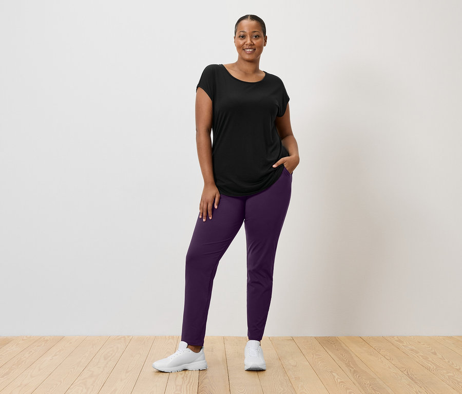 Frau mit schwarzem T-Shirt, lila Hose und weißen Turnschuhen steht vor einer weißen Wand.