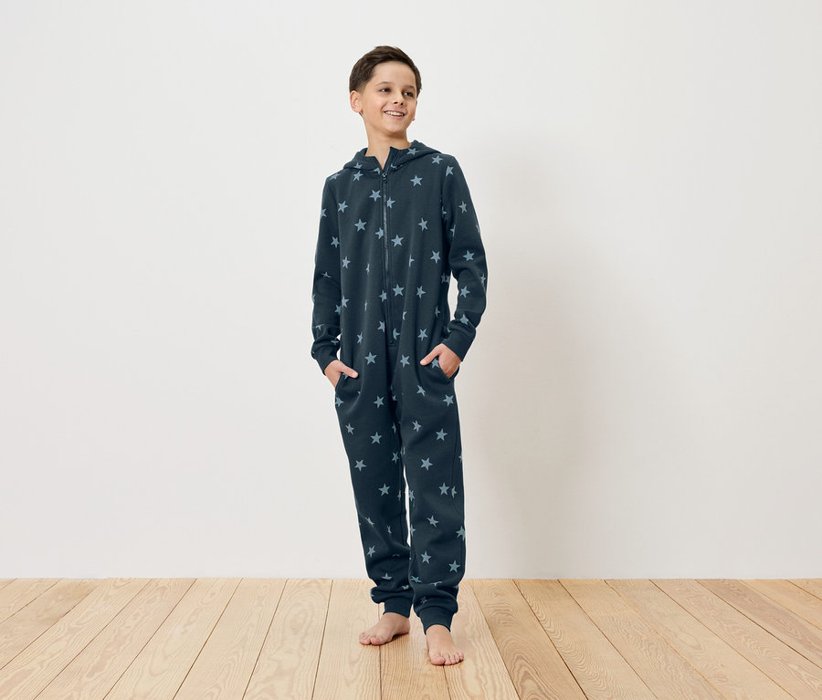 Ein junger Mann steht barfuß auf einem Holzboden, er trägt einen dunkelblauen Sternen-Overall mit Kapuze und Reißverschluss und hat die Hände in den Taschen.