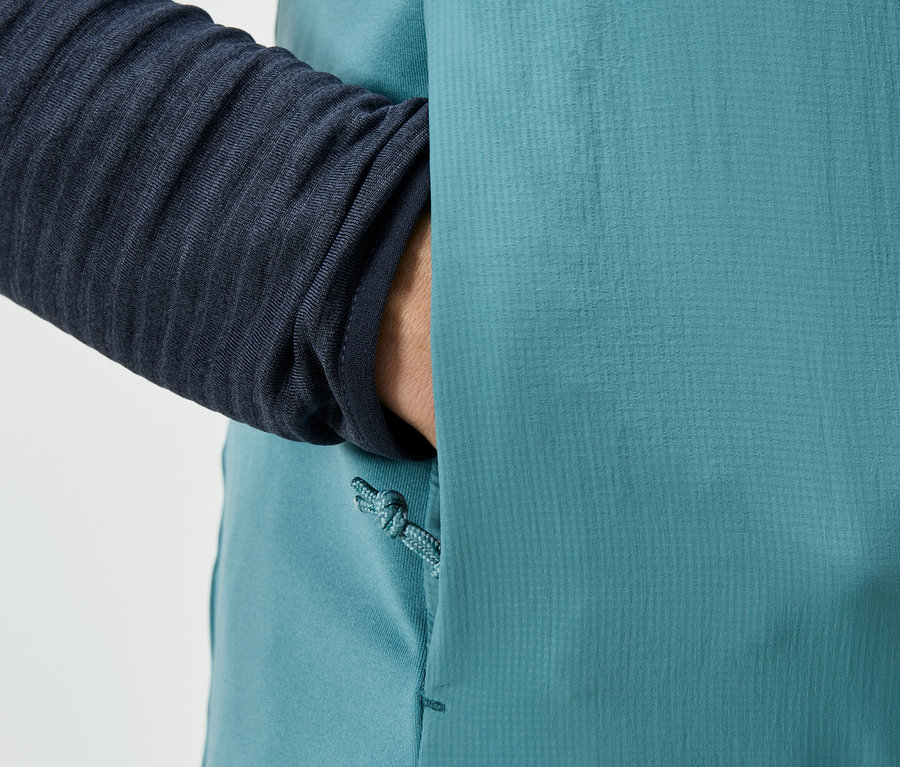 Detailaufnahme einer Person, die eine Hand in die Tasche einer türkisfarbenen Jacke steckt, mit einem marineblauen Ärmel sichtbar.