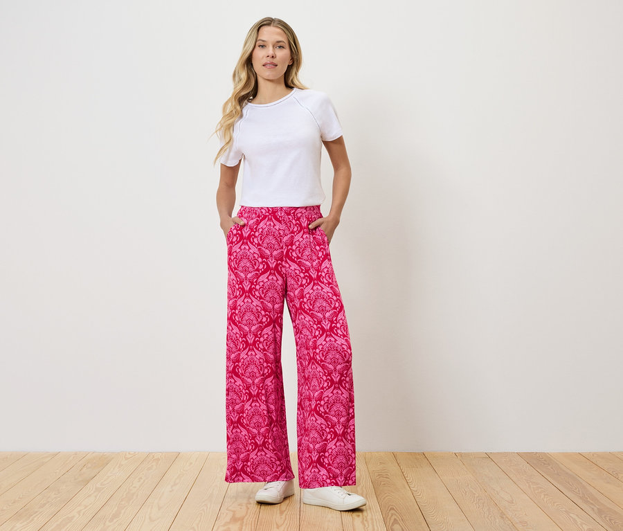 Frau steht mit Händen in den Taschen einer pinkfarbenen Hose mit Muster.