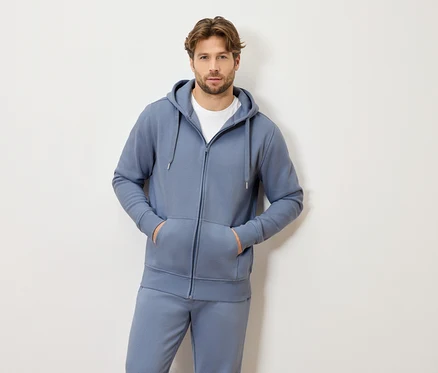 Mann mit hellbraunem Haar, der einen blauen Trainingsanzug mit Kapuze und Reißverschluss und ein weißes T-Shirt trägt und mit beiden Händen in den Taschen steht.