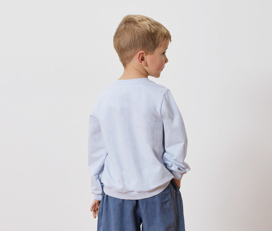 Ein blonder Junge steht mit dem Rücken zur Kamera, er trägt ein hellblaues Sweatshirt und blaue Hosen.