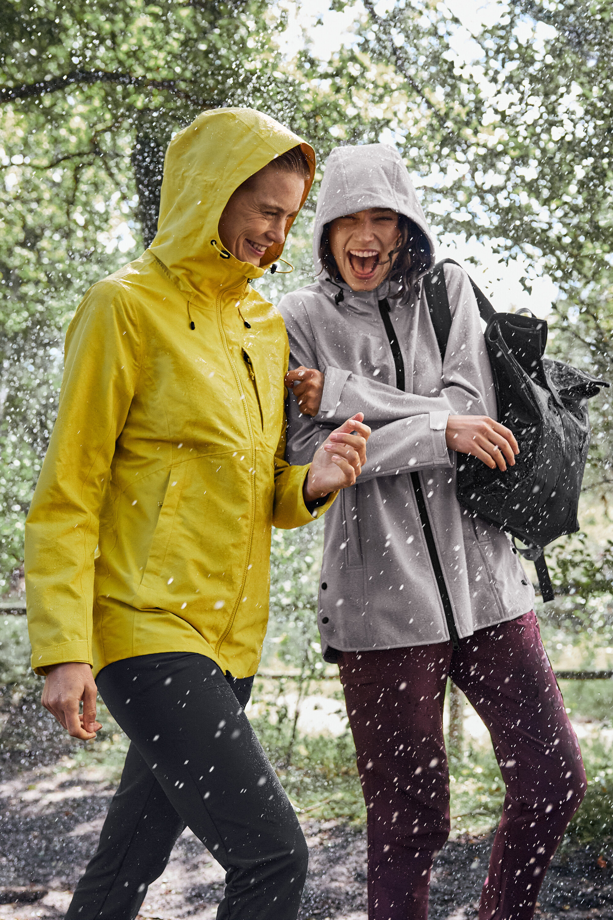 Zwei Frauen gehen im Regen, die eine trägt eine gelbe Regenjacke, die andere eine graue mit einem schwarzen Rucksack.