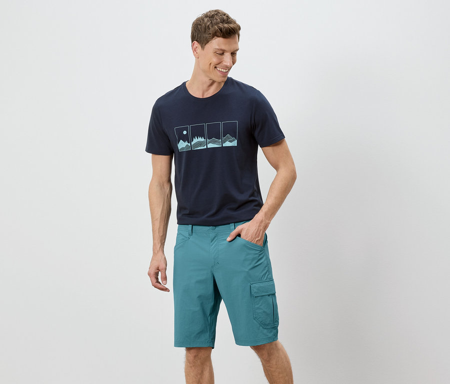 Ein Mann steht mit einer Hand in der Tasche vor einem weißen Hintergrund. Er trägt ein marineblaues T-Shirt mit einer Grafik und grüne Shorts.