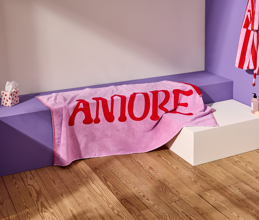 Ein rosa Handtuch mit dem roten Wort "AMORE" liegt auf einer violetten Bank. Ein gestreifter Bademantel hängt an der Wand.