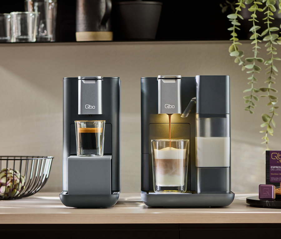 Zwei dunkelgraue Qbo MILK MASTER Kaffeemaschinen auf einer Theke. Eine Maschine hat einen Espresso, die andere bereitet einen Latte Macchiato zu. Daneben liegen Kaffeekapseln.