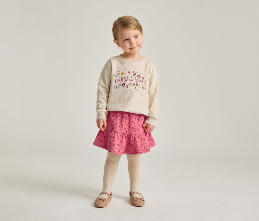Ein kleines Mädchen mit blonden Haaren trägt ein beiges Sweatshirt mit der Aufschrift "Wild and loved", einen rosa Rock und Ballerinaschuhe.