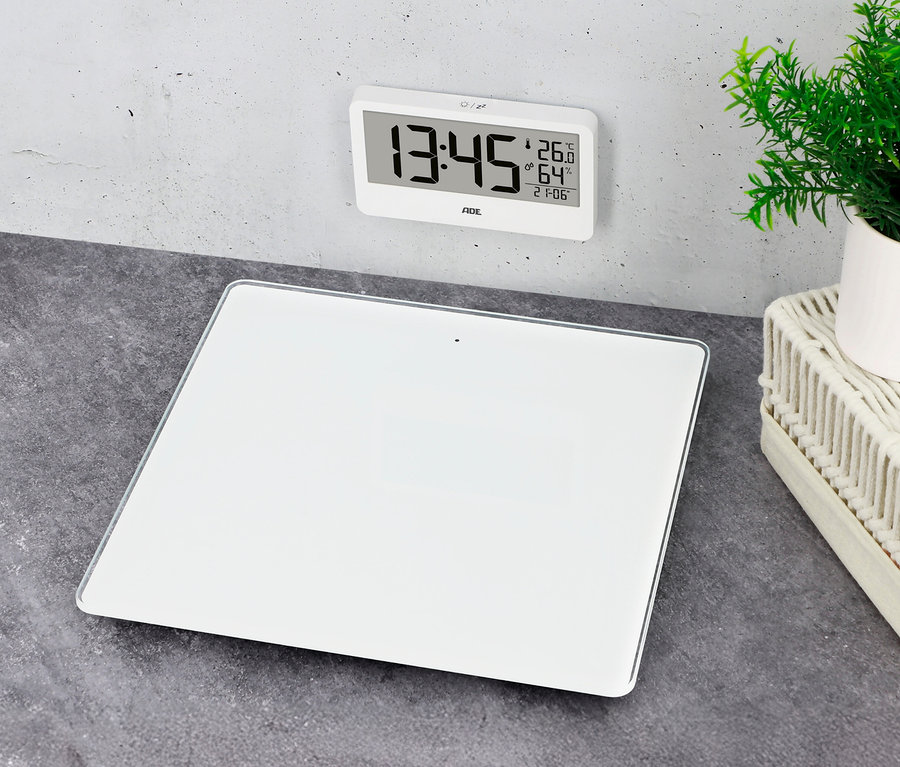 Weiße digitale Personenwaage auf dunklem Untergrund. Darüber hängt eine weiße Digitaluhr, die Zeit und Temperatur anzeigt. Rechts steht eine kleine Pflanze in einem weißen Topf.