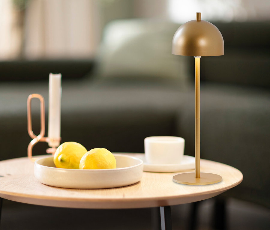 Goldfarbene SCHÖNER WOHNEN Akku-Tischleuchte »Bellis« auf einem Holztisch, daneben eine Schale mit Zitronen, eine Kerze und eine Tasse.