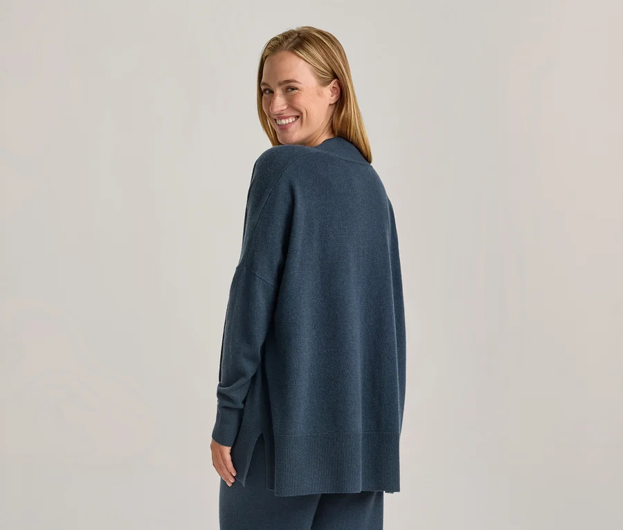Frau in einem blauen, locker sitzenden Pullover und passender Hose, die über ihre Schulter blickt und lächelt.