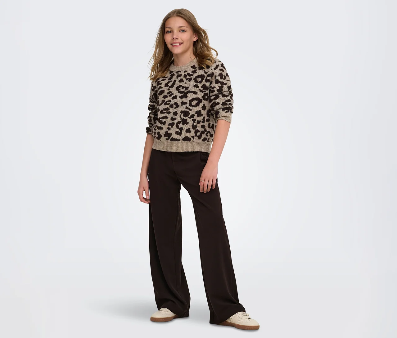 Ein junges Mädchen steht lächelnd da und trägt einen beigefarbenen Pullover mit Leopardenmuster, eine dunkelbraune weite Hose und helle Sneaker.