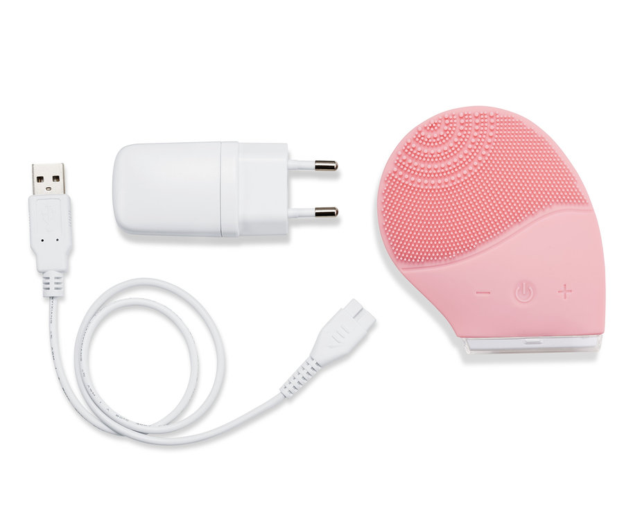 Eine elektrische Silikon-Gesichtsreinigungsbürste in Rosa liegt mit Ladekabel und Adapter auf weißem Grund.