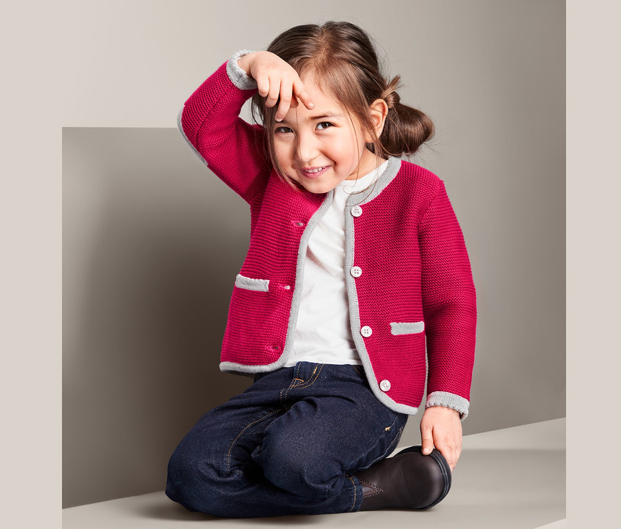 Ein Mädchen kniet und trägt eine Strickjacke im Trachtenlook, Jeans und Stiefel.