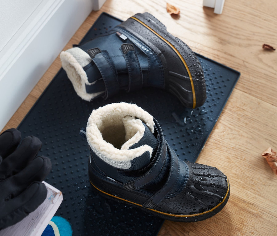 Zwei Allwetterstiefel liegen auf einer dunkelblauen Fußmatte neben einer weißen Tür.