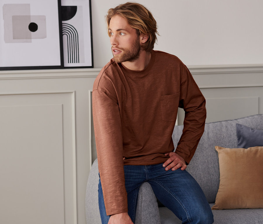Mann sitzt auf grauem Sofa, trägt ein braunes 2 Langarmshirts mit Baumwolle aus nachhaltigen Quellen und Jeans.